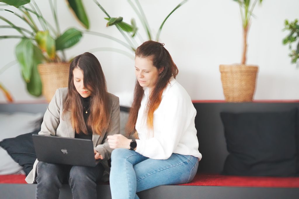 Zwei Frauen sitzen auf einer Bank und schauen gemeinsam auf einen Laptop.