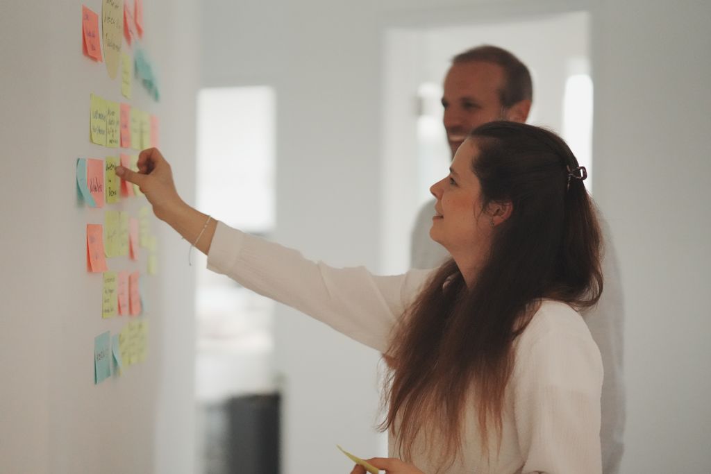 Eine Frau klebt eine Notiz an eine Wand voller bunter Post-its, ein Mann lächelt im Hintergrund.
